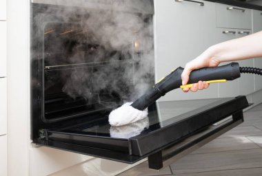 Kadın eli fırının cam kapısını temizlemek için buhar temizleyici kullanıyor. Çevre dostu mutfak temizliği.