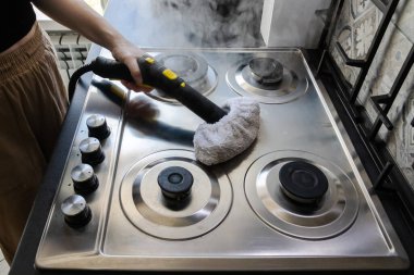 Kadın elinin, paslanmaz çelik gaz sobasından yağ ve kalıntı çıkarmak için buhar temizleyicisi kullanması. Çevre dostu mutfak temizliği.