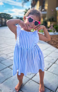 Güneşli bir yaz gününde kalp şeklinde güneş gözlüklü gülümseyen kız. Mavi beyaz çizgili bir elbise içinde kameraya poz vermek. Yalınayak 