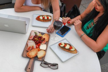 Friends enjoying a chic brunch outdoors with a glass of red wine, charcuterie board and sandwiches, accompanied by a laptop and smartphone, personifying modern gastronomy. clipart