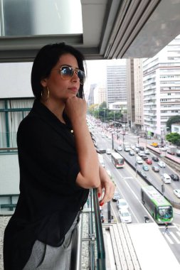 A woman wearing sunglasses stands on a balcony overlooking a busy city street with traffic and tall buildings, reflecting a sense of contemplation and modern urban lifestyle. clipart