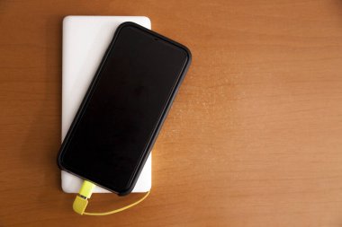 Siyah akıllı telefon, beyaz bir güç bankası tarafından ahşap bir masanın üstünde şarj ediliyor. Güç bankası konsepti. Teknoloji konsepti.