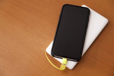 Siyah akıllı telefon, beyaz bir güç bankası tarafından ahşap bir masanın üstünde şarj ediliyor. Güç bankası konsepti. Teknoloji konsepti.