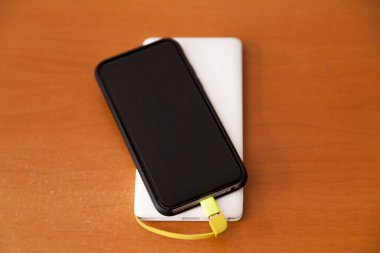 Siyah akıllı telefon, beyaz bir güç bankası tarafından ahşap bir masanın üstünde şarj ediliyor. Güç bankası konsepti. Teknoloji konsepti.