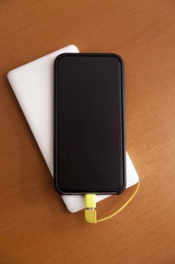 Siyah akıllı telefon, beyaz bir güç bankası tarafından ahşap bir masanın üstünde şarj ediliyor. Güç bankası konsepti. Teknoloji konsepti.