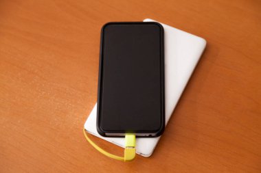 Siyah akıllı telefon, beyaz bir güç bankası tarafından ahşap bir masanın üstünde şarj ediliyor. Güç bankası konsepti. Teknoloji konsepti.