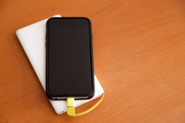 Siyah akıllı telefon, beyaz bir güç bankası tarafından ahşap bir masanın üstünde şarj ediliyor. Güç bankası konsepti. Teknoloji konsepti.