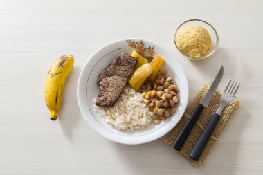 Bir tabak pilav, fasulye, biftek ve manyok. Çatal bıçak takımı, manyok unu ve tatlı olarak da muz. Brezilya mutfağında beyaz ahşap arka plan, üst manzara. Yemek konsepti. Makarna konsepti.