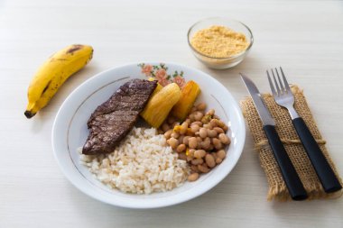 Bir tabak pilav, fasulye, biftek ve manyok. Çatal bıçak takımı, manyok unu ve tatlı olarak da muz. Brezilya mutfağında beyaz ahşap arka plan, üst manzara. Yemek konsepti. Makarna konsepti.