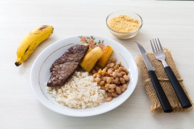 Bir tabak pilav, fasulye, biftek ve manyok. Çatal bıçak takımı, manyok unu ve tatlı olarak da muz. Brezilya mutfağında beyaz ahşap arka plan, üst manzara. Yemek konsepti. Makarna konsepti.