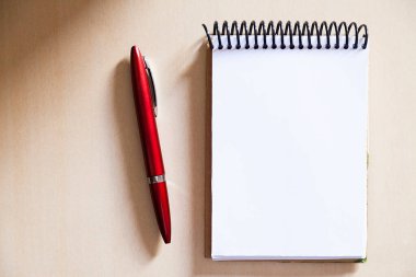 Tahta masanın üzerinde boş bir not defteri, yanında kırmızı bir kalem, yanına da güneş ışığı vuruyor. Hatırlatmalar için not defteri. Açıklama konsepti. Nesne konsepti. Boşluğu kopyala