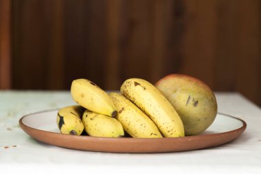 Meyveler masanın üzerindeki bir tabağa konur. Meyveler muz ve mangodur. Brezilya mutfağında ahşap kaplama. Uzayı kopyala, üst görünüm. Yemek konsepti. Meyve konsepti.