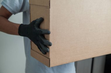 Gri arkaplandaki ürünleri teslim etmek için elinde karton bir kutu tutan siyah eldivenli adamın ayrıntıları. Teslimat konsepti. Teslimat hizmeti konsepti. Boşluğu kopyala.