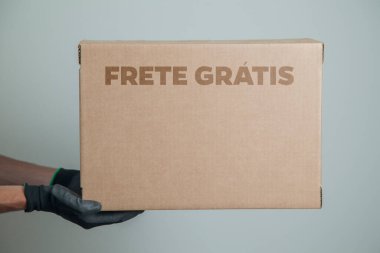 Elinde karton bir kutu tutan siyah eldivenli bir adamın ayrıntıları. Gri arka planda ürünleri teslim etmek için ücretsiz kargo. Teslimat konsepti. Teslimat hizmeti konsepti. Uzayı kopyala Kara Cuma konsepti.