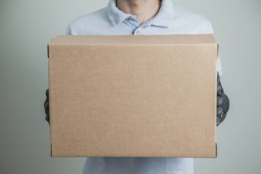 Gri arkaplandaki ürünleri teslim etmek için elinde karton bir kutu tutan siyah eldivenli adamın ayrıntıları. Teslimat konsepti. Teslimat hizmeti konsepti. Boşluğu kopyala.