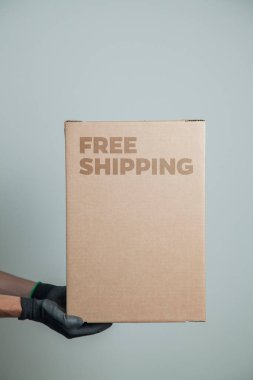 Elinde karton bir kutu tutan siyah eldivenli bir adamın ayrıntıları. Gri arka planda ürünleri teslim etmek için ücretsiz kargo. Teslimat konsepti. Teslimat hizmeti konsepti. Uzayı kopyala Kara Cuma konsepti.