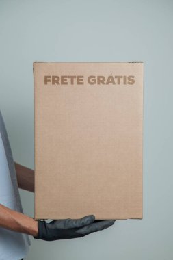 Elinde karton bir kutu tutan siyah eldivenli bir adamın ayrıntıları. Gri arka planda ürünleri teslim etmek için ücretsiz kargo. Teslimat konsepti. Teslimat hizmeti konsepti. Uzayı kopyala Kara Cuma konsepti.