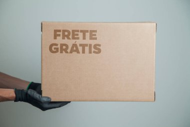 Elinde karton bir kutu tutan siyah eldivenli bir adamın ayrıntıları. Gri arka planda ürünleri teslim etmek için ücretsiz kargo. Teslimat konsepti. Teslimat hizmeti konsepti. Uzayı kopyala Kara Cuma konsepti.