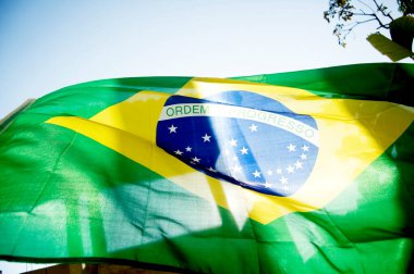 Brezilya bayrağı dalgalanıyor. Arka planda mavi gökyüzü ve güneş parlıyor.