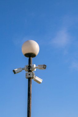 Yuvarlak beyaz ampullü bir sokak lambası, birden fazla güvenlik kamerasıyla çevrili bir direğe monte edilir. Açık mavi gökyüzü, güneşli bir günü işaret eden bir zemin görevi görür..