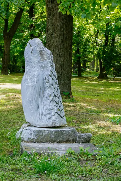 Taştan yapılmış heykel, huzurlu bir parktaki yemyeşil ağaçların arasında duruyor..
