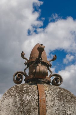 Karmaşık bir metal heykel yıpranmış bir kubbeyi taçlandırıyor, özenle hazırlanmış tasarımlar ve pas sergiliyor. Kabarık bulutlarla karşıt mavi gökyüzü bu mimari detay için çarpıcı bir zemin oluşturur..