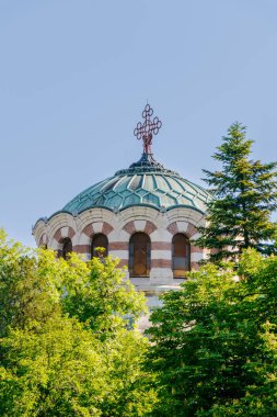 Tarihi bir kilisenin karmaşık kubbesi berrak mavi gökyüzünün altındaki canlı ağaçların arasında duruyor. Detaylı mimari özellikler ve gündüz boyunca huzurlu bir atmosfer sergiliyor..