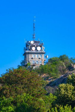 Uydu anteni ve antenlerle donatılmış yüksek bir telekomünikasyon kulesi yamaçta belirgin bir şekilde duruyor. Yapıyı çevreleyen yemyeşil ağaçlar, gökyüzü parlak ve berrak kalır..