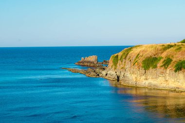 Kayalık bir kıyı şeridinin altın saat boyunca sakin mavi sularla buluşması nefes kesici bir manzaraydı. Dalgalar kayalıklara çarpıyordu. Açık gökyüzü sahnenin doğal güzelliğini arttırır..