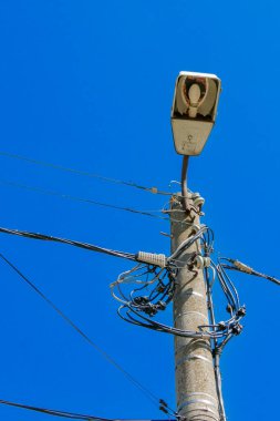 Bir elektrik direği, açık mavi bir gökyüzüne karşı bir sokak lambasını ve sayısız elektrik kablosunu destekleyen uzun bir duruş sergiliyor. Açık gün, kutbun ve bileşenlerinin detaylarını vurgular..