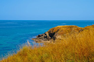 Açık bir yaz gününde Karadeniz 'in parlak mavi sularına bakan bir kıyı kayalığının panoramik manzarası. Altın kuru otlar ön plandaki yamaçları kaplar..