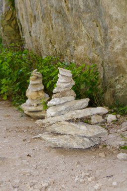 İki dikkatlice istiflenmiş taş oluşumu yeşilliklerle çevrili kayalık bir duvarın yanında otururlar..