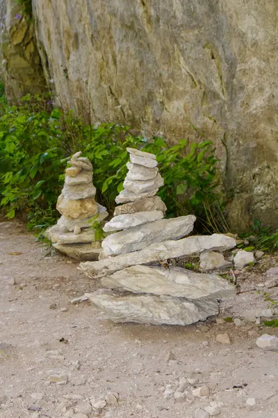 İki dikkatlice istiflenmiş taş oluşumu yeşilliklerle çevrili kayalık bir duvarın yanında otururlar..