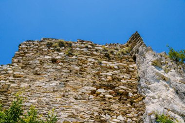 Ortaçağ duvarının yıpranmış taş kalıntıları, yaz mevsiminde bitki örtüsü ve mavi gökyüzü tarafından çerçevelenmiş bir dağın kayalık arazisinde birleşir..
