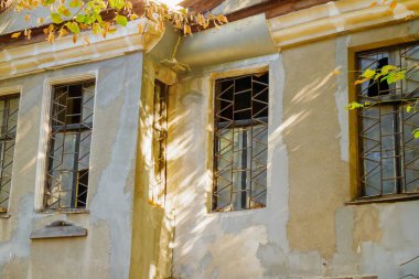 Yaşlanmış bir şehir binası çatlak alçı ve parmaklıklı camlarla aşınma izleri gösterirken, yumuşak güneş ışığı desenli duvarların üzerindeki yapraklardan süzülüyor..