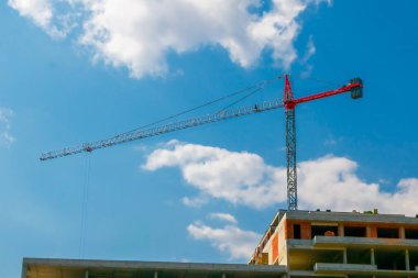 Güneşli bir günde kısmen inşa edilmiş bir binanın üzerinde yüksek bir vinç kulesi, açık mavi gökyüzü ve dağınık bulutlara karşı devam eden gelişim çalışmalarını sergiliyor.