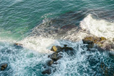 Gün batımında kıyı şeridi boyunca kayalara çarpan canlı dalgalar hafif dalgalar ve zengin renklerle deniz kenarında doğanın gücünün ve güzelliğinin çarpıcı bir görüntüsünü oluşturur..