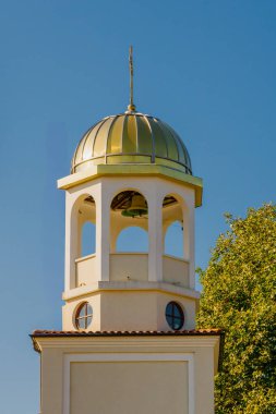 Eşsiz çan kulesi, üzerinde parlak mavi gökyüzü ve yakınındaki canlı yeşil ağaçlarla vurgulanmış altın bir kubbeyle ayakta duruyor. Çekicilikle dolu güzel bir gündüz ortamı yaratıyor..