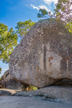 Dev bir granit kaya, orman ağaçları ve açık havada mavi gökyüzü ile çevrili daha küçük kayalar üzerinde mükemmel bir şekilde dengelenmiş görünüyor..