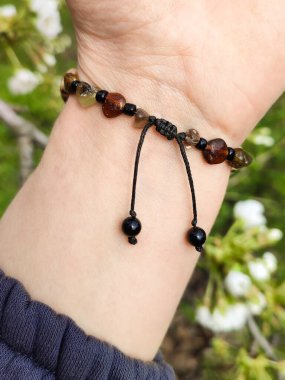 Bileğinde hakiki kehribar olan kahverengi boncuklu bir bilezik. Makrame bilezik kayması düğümlenmiş kapanış. Çiçekler arka planda. Bahar zamanı, güneş ışığı. Aksesuar, moda, tarz, mücevher. 