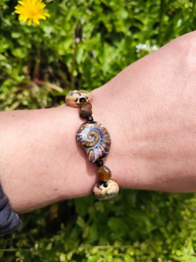 Kahverengi boncuklu etnik tarzda bilezik, hakiki kehribar, salyangoz şeklinde seramik boncuklar kadının elinde. Yeşil çimenler, arka planda karahindiba. Bahar zamanı, güneş ışığı. Aksesuar, moda, tarz, mücevher.