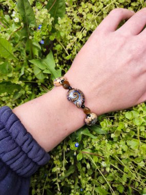 Kahverengi boncuklu etnik tarzda bilezik, hakiki kehribar, salyangoz şeklinde seramik boncuklar kadının elinde. Yeşil çimenler, arka planda karahindiba. Bahar zamanı, güneş ışığı. Aksesuar, moda, tarz, mücevher.