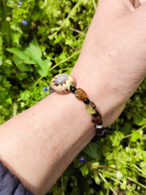 Kahverengi boncuklu etnik tarzda bilezik, hakiki kehribar, kadının elinde seramik boncuklar. Yeşil çimenler, arka planda karahindiba. Bahar zamanı, güneş ışığı. Aksesuar, moda, tarz, mücevher.