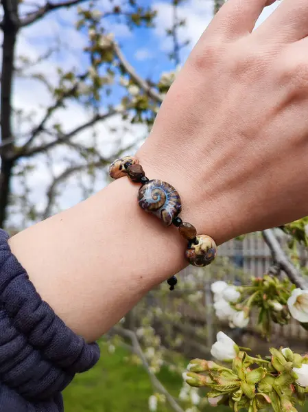 Kahverengi boncuklu etnik tarzda bilezik, hakiki kehribar, salyangoz şeklinde seramik boncuklar kadının elinde. Mavi gökyüzü, arka planda beyaz bahar çiçekleri, gün ışığı. Doğa ilham verici moda aksesuarı. 