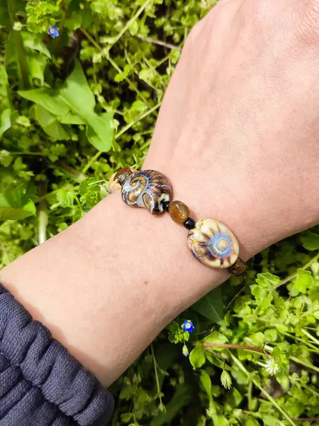 Kahverengi boncuklu etnik tarzda bilezik, hakiki kehribar, salyangoz şeklinde seramik boncuklar kadının elinde. Yeşil çimenler, arka planda karahindiba. Bahar zamanı, güneş ışığı. Aksesuar, moda, tarz, mücevher.