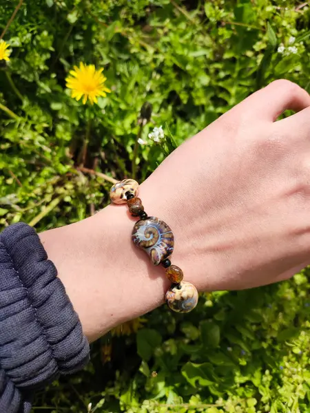 Kahverengi boncuklu etnik tarzda bilezik, hakiki kehribar, salyangoz şeklinde seramik boncuklar kadının elinde. Yeşil çimenler, arka planda karahindiba. Bahar zamanı, güneş ışığı. Aksesuar, moda, tarz, mücevher.