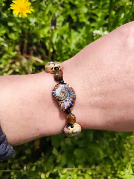 Kahverengi boncuklu etnik tarzda bilezik, hakiki kehribar, salyangoz şeklinde seramik boncuklar kadının elinde. Yeşil çimenler, arka planda karahindiba. Bahar zamanı, güneş ışığı. Aksesuar, moda, tarz, mücevher.