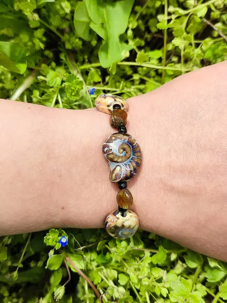 Kahverengi boncuklu etnik tarzda bilezik, hakiki kehribar, salyangoz şeklinde seramik boncuklar kadının elinde. Yeşil çimenler, arka planda karahindiba. Bahar zamanı, güneş ışığı. Aksesuar, moda, tarz, mücevher.