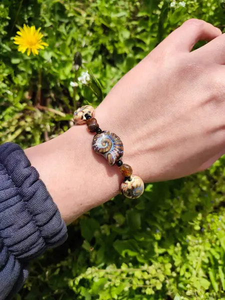 Kahverengi boncuklu etnik tarzda bilezik, hakiki kehribar, salyangoz şeklinde seramik boncuklar kadının elinde. Yeşil çimenler, arka planda karahindiba. Bahar zamanı, güneş ışığı. Aksesuar, moda, tarz, mücevher.