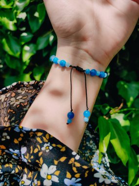 Mavi taşlar, kadının bileğindeki kyanit apatit boncuklu bilezik. Yeşil yapraklar arka plan, güneş ışığı, gölgeler, yaz. Yarı değerli taş makrome ayarlanabilir kilit bilezik, mücevher, aksesuar. 